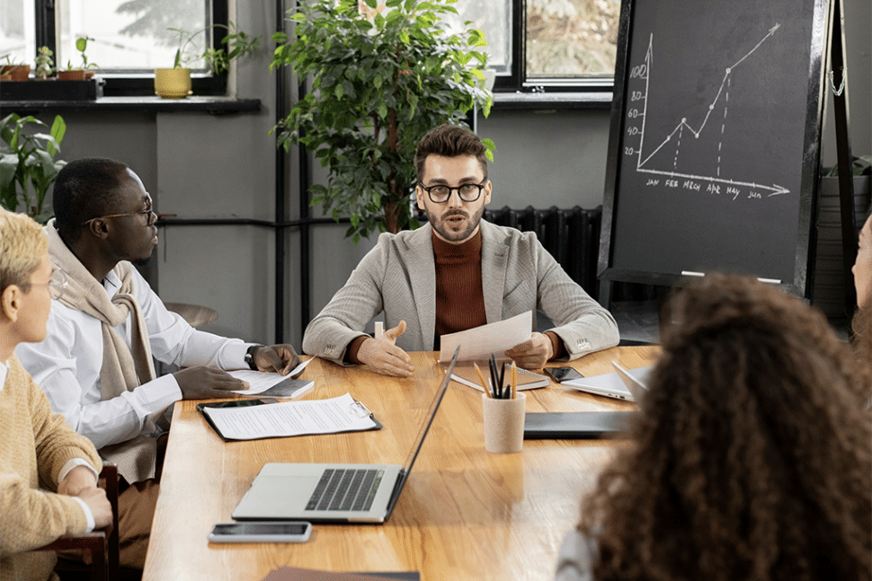 Четыре человека встречаются в комнате с ноутбуком, бумагами, ручками и доской.