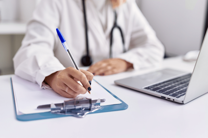 A doctor writes some notes on a clipboard, sitting at a desk with an open laptop